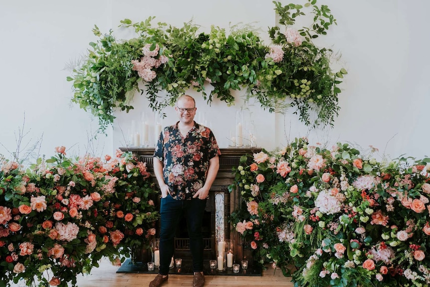 A man in a floral shirt stands in front of a large green and pink floral arrangement