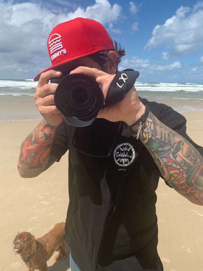 A man wearing a black t-shirt and red cat holds a camera on a beach.