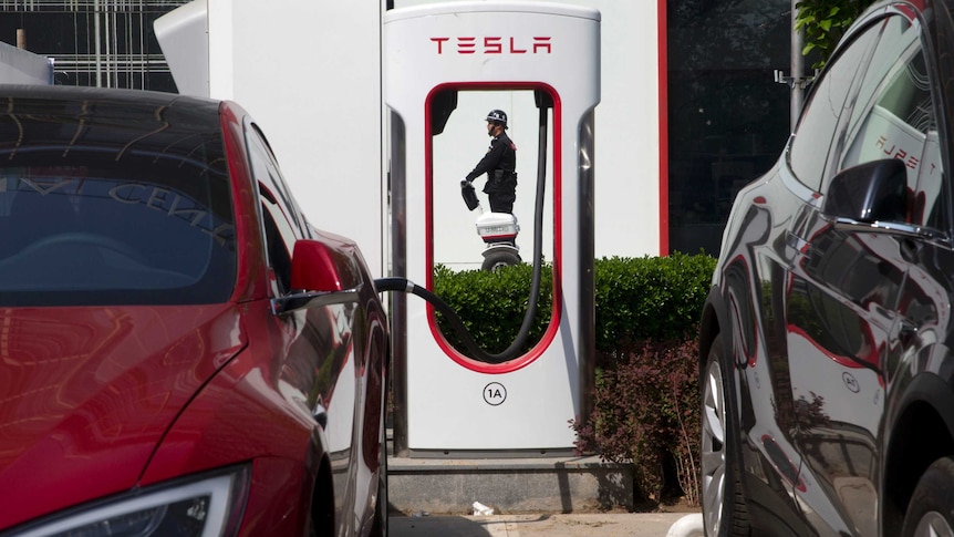 A red electric Tesla vehicle charges while parked next to a black Tesla as security guard is seen walking in the background.