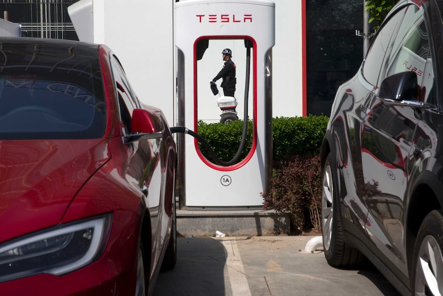 A red electric Tesla vehicle charges while parked next to a black Tesla as security guard is seen walking in the background.