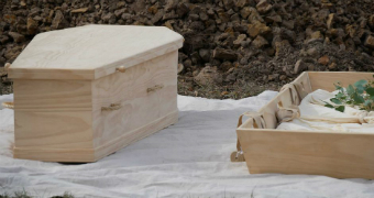 An untreated wooden coffin next to an open shallow rectangular box with a shroud in it.