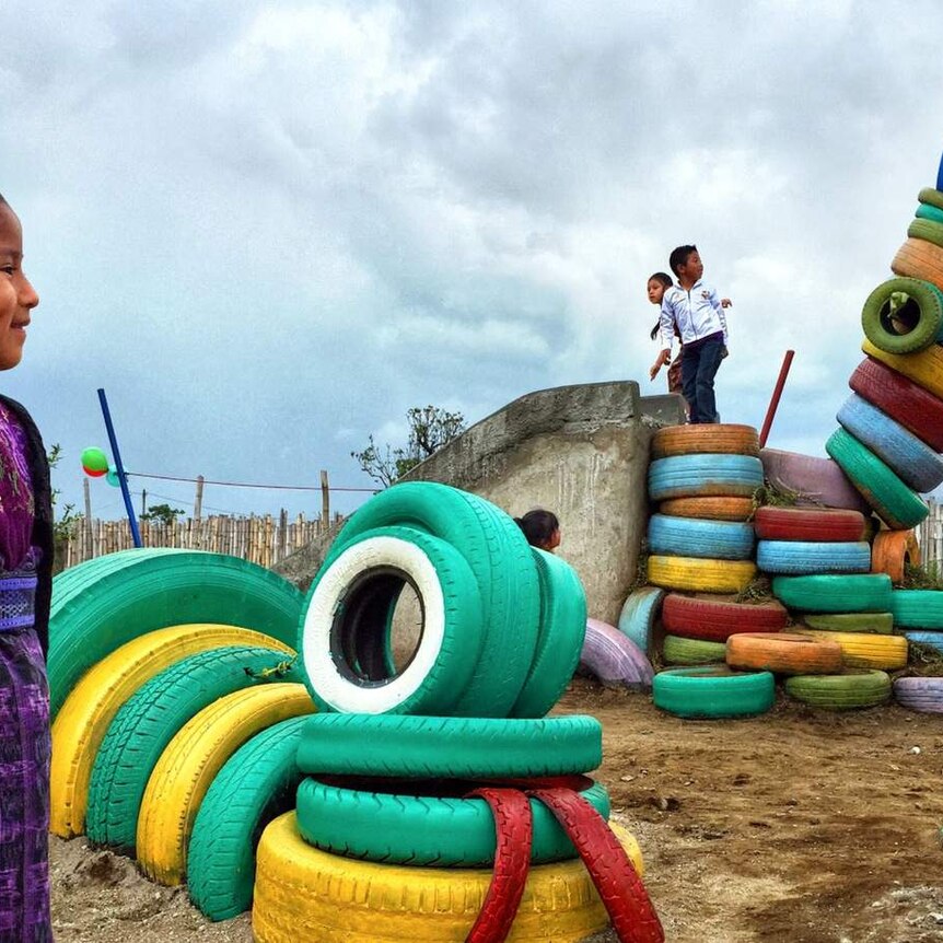 A young girl stands smiling in the foreground as she watches other children play on a dinosaur made of tires.