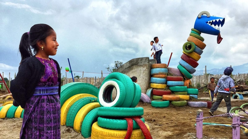 A young girl stands smiling in the foreground as she watches other children play on a dinosaur made of tires.