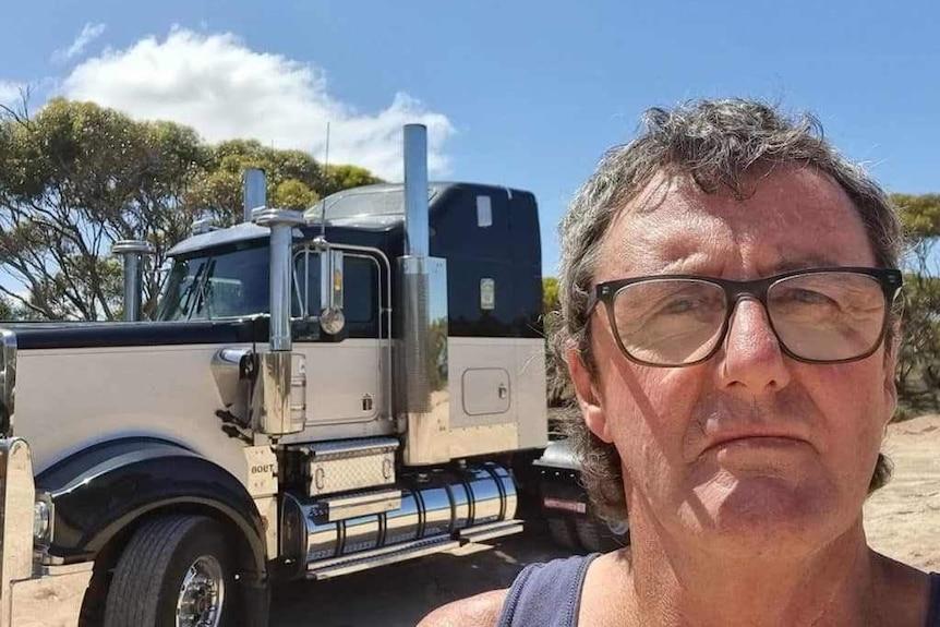 A bespectacled man in a singlet stands near a truck.