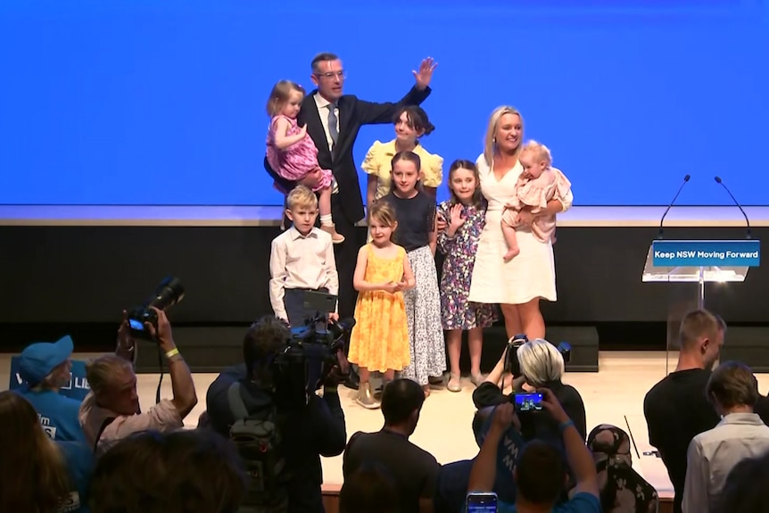 a man wearing glasses standing on a stage holding a young  girl he's surrounded by a woman and six other children