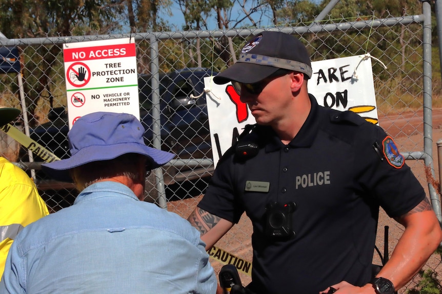 a police officer talks to a man in a blue shirt and hat in front of mesh fencing