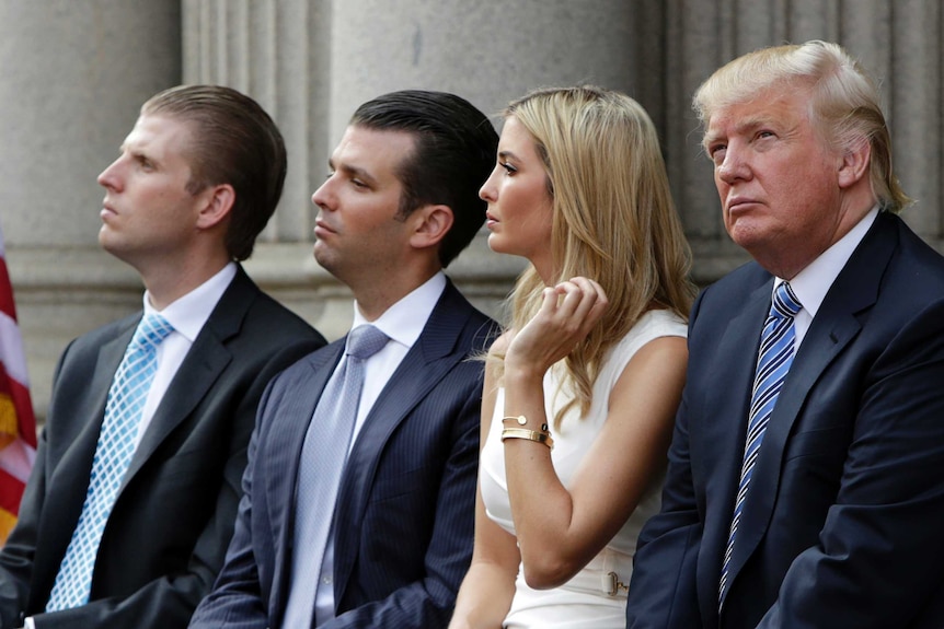 Three men in navy suits and pale blue ties sit in front of a official building with a blonde woman in a white, sleeveless dress