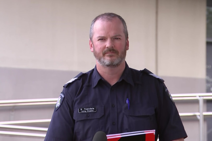 The police officer wears a navy police shirt with his name tag and a pen in his pocket and holds a red and black notebook.