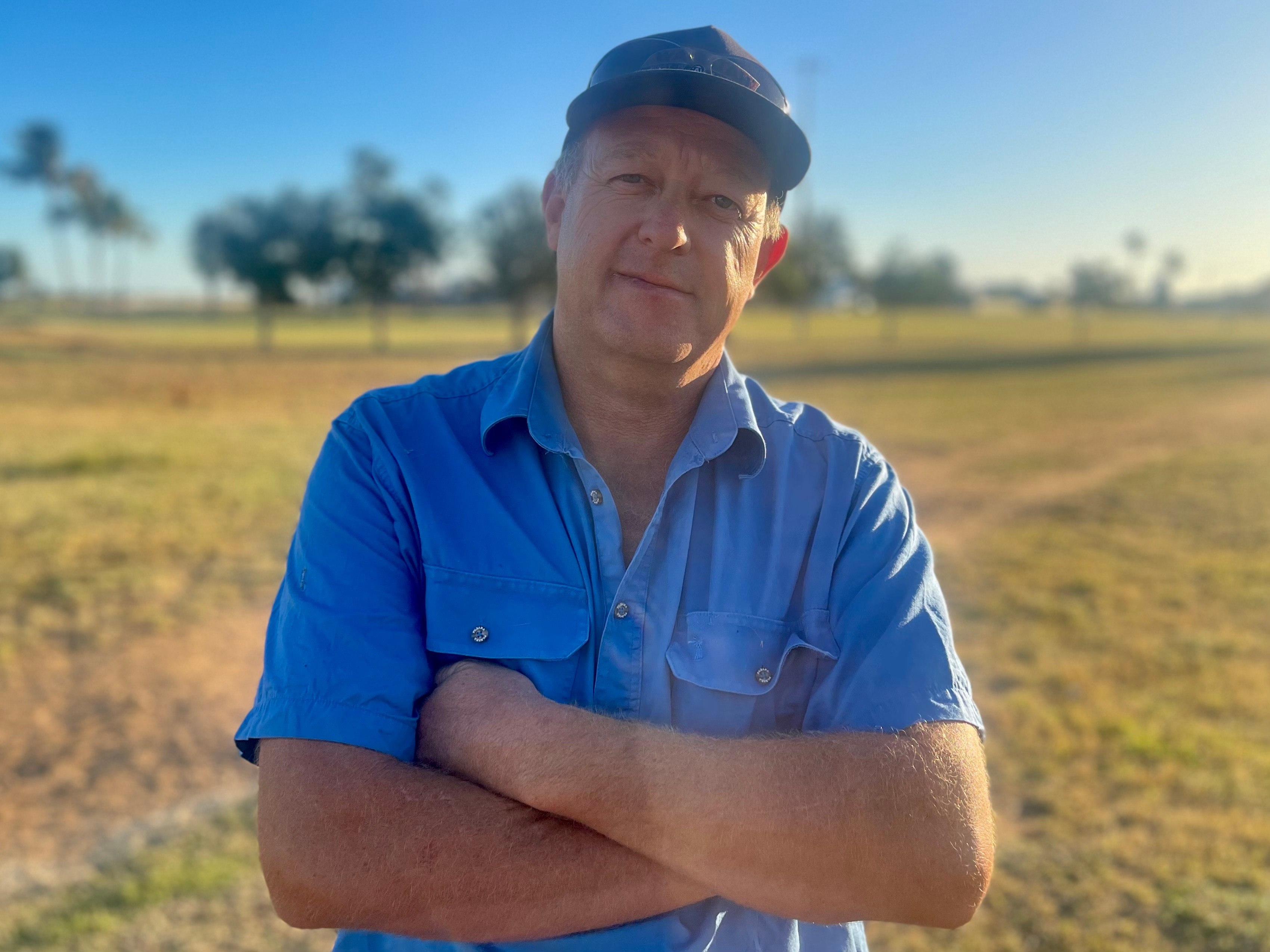 A pastoralist stands with his arms crossed and a serious face in a blue shirt and cap. 