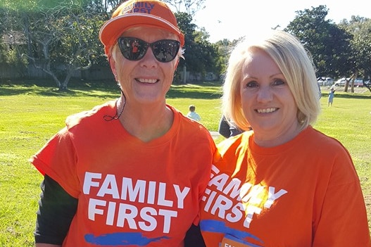 Two women stand in orange family first shirts holding how to vote cards.