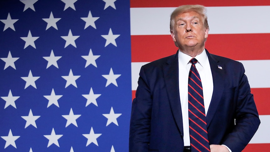 Donald Trump stands holding his belt buckle while in front of a US flag