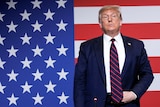 Donald Trump stands holding his belt buckle while in front of a US flag