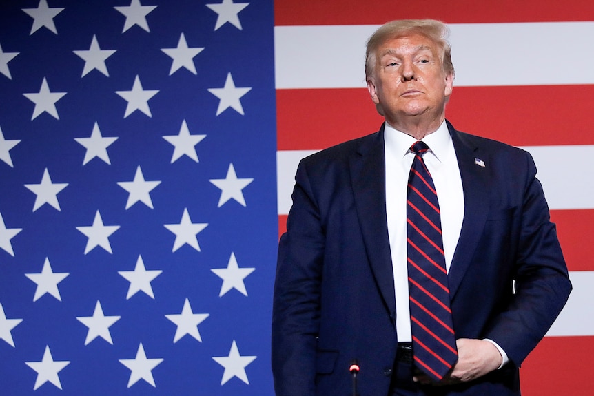 Donald Trump stands holding his belt buckle while in front of a US flag