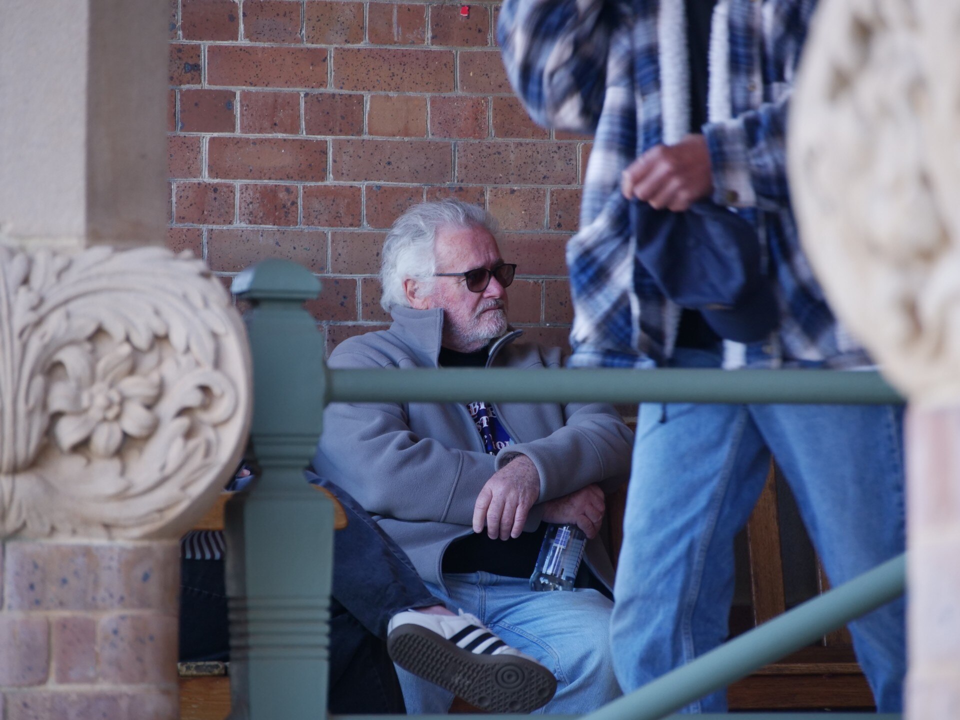 A man with grey hair and sunglasses sitting down with a person walking past.