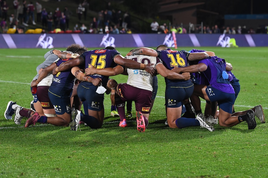NRL players kneel in a circle, with their arms around each other as they pray.