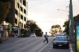 Woman wearing a mask crosses a street at Brisbane's West End at sunset.