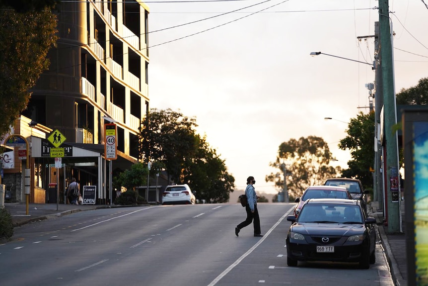 Woman wearing a mask crosses a street at Brisbane's West End at sunset.