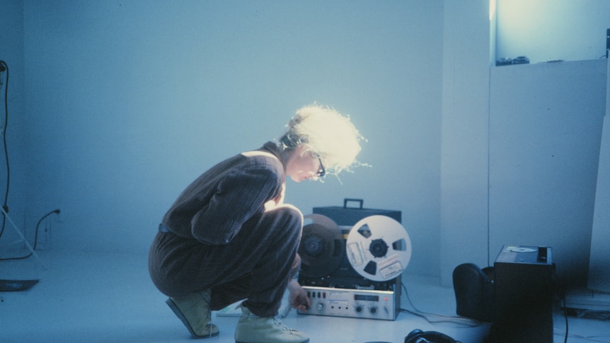 Photo of woman with light hair tied up, crouching over transistor in bare room with bluish light.