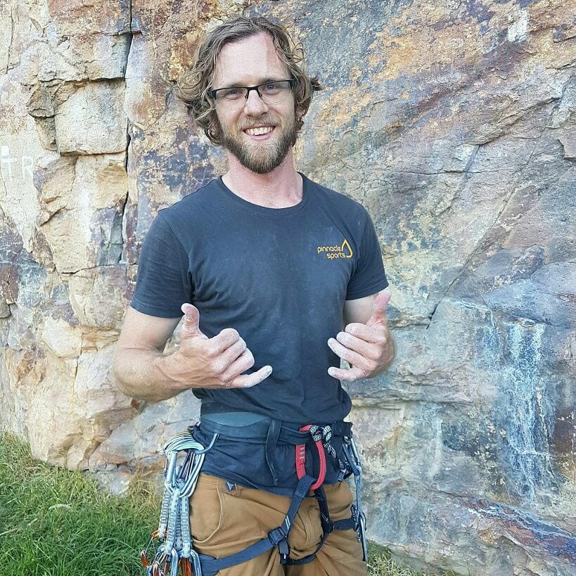 Josh Worley in a rock climbing harness standing in front of a rock wall.