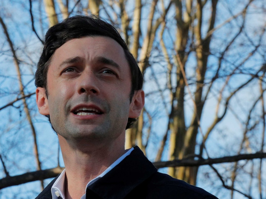 A white man with brown hair looking up and in the middle of a sentence with bare trees behind him