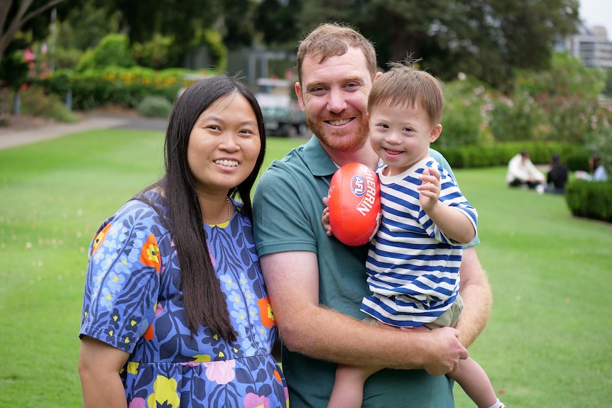 Keiran holds young Henry in his arms, who is holding a small football. Jenny stands next to them.