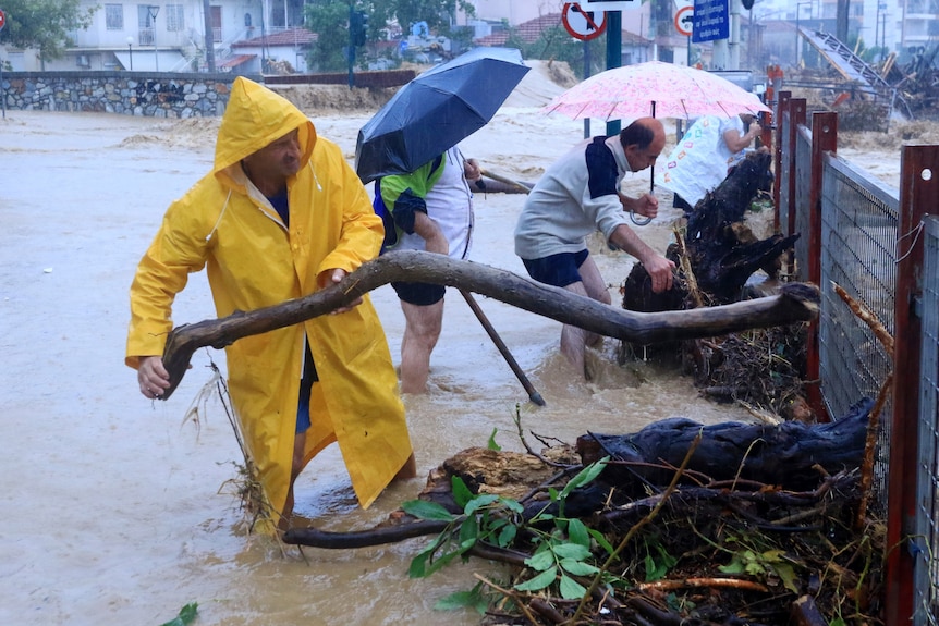 Un hombre vestido con un impermeable amarillo levanta una rama del agua hasta las pantorrillas. 