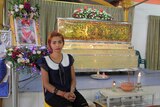 A young woman sits in front of a coffin, next to the framed photo of a baby girl.