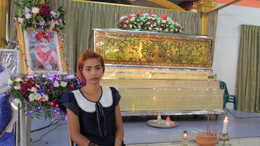 A young woman sits in front of a coffin, next to the framed photo of a baby girl.