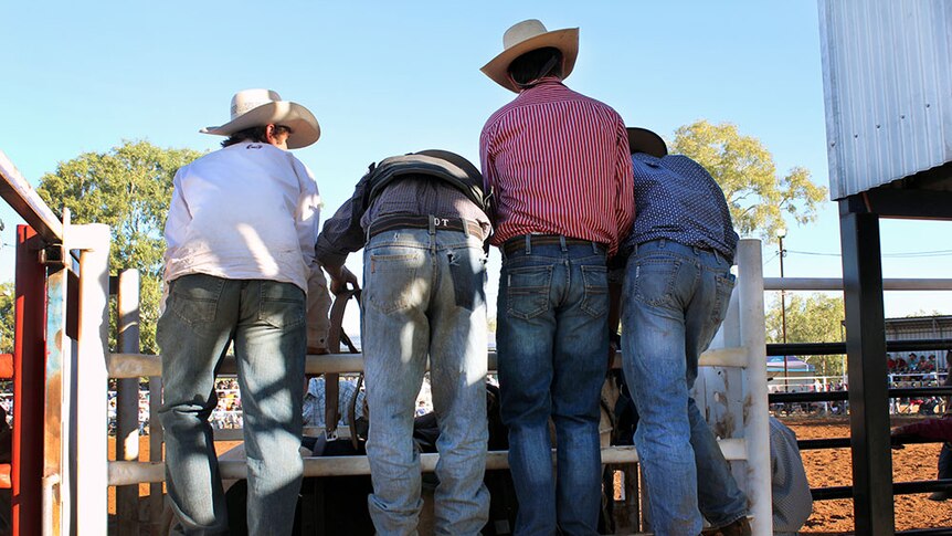 Four men dressed in cowboy hats, boots, and blue jeans, lean over to help a friend in the shoots.