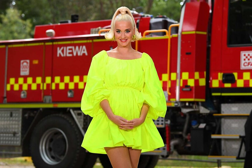 Katy Perry wears a florescent yellow dress and stands in front of a fire truck with hands clasped in front of her.