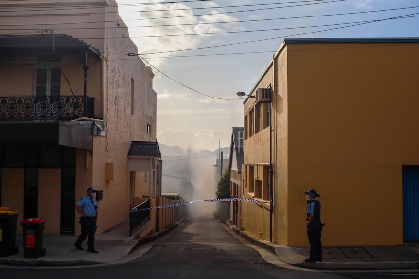 Two police stand on either side of a closed-off laneway with smoke hanging in the air and a police tape between two buildings