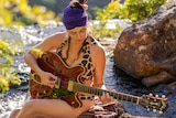 Sophia Fletcher playing electric guitar while sitting on rocks alongside a stream.