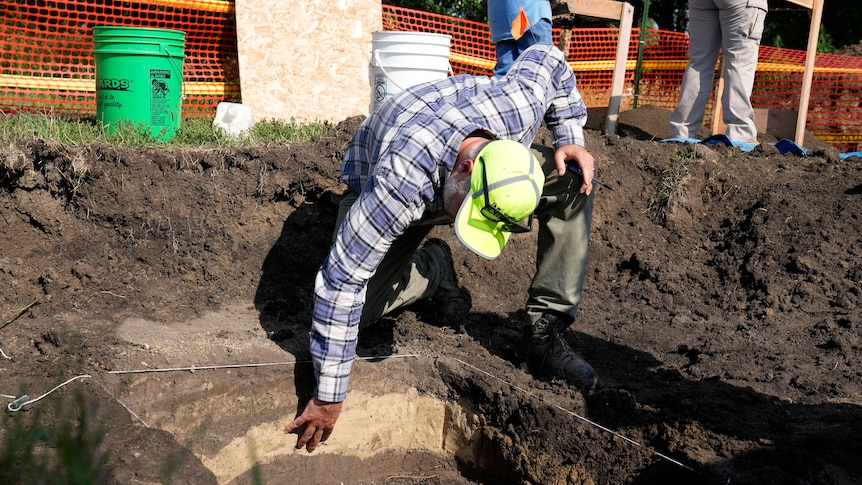 A man in a fleuro hat bends over and reaches his hand down into layers of excavated soil. 