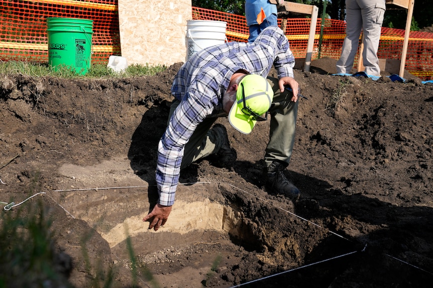 A man in a fleuro hat bends over and reaches his hand down into layers of excavated soil. 