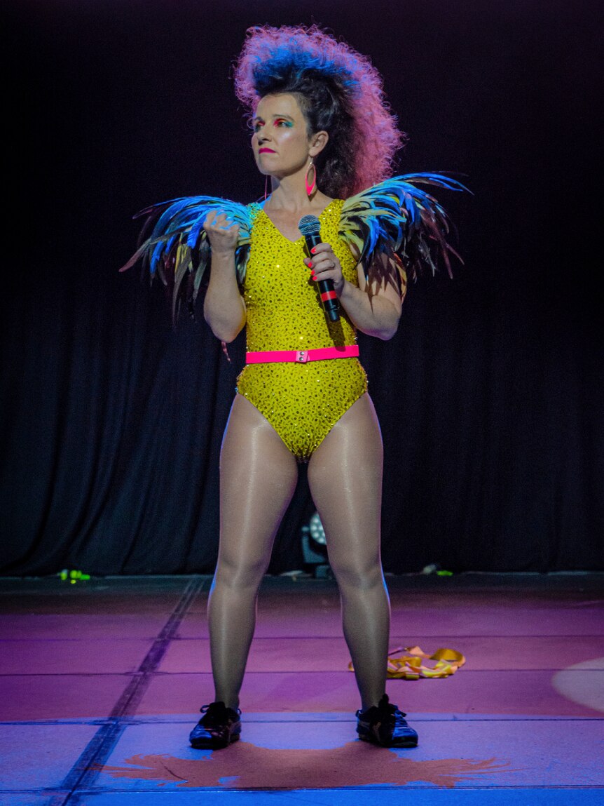 Woman with large teased hair and yellow leotard stands on stage
