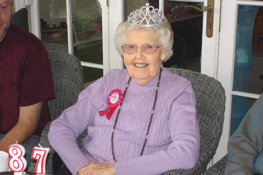 Audrey MacGregor with a 'birthday girl' badge and tiara on her head, sitting in front of a cake with an '87' birthday candle.