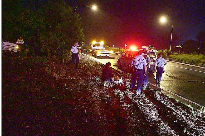 Two people sit by the side of the road as police look at the scene of a car crash