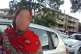 A woman wearing a headscarf stands in front of her car which has been pulled over by a police car.