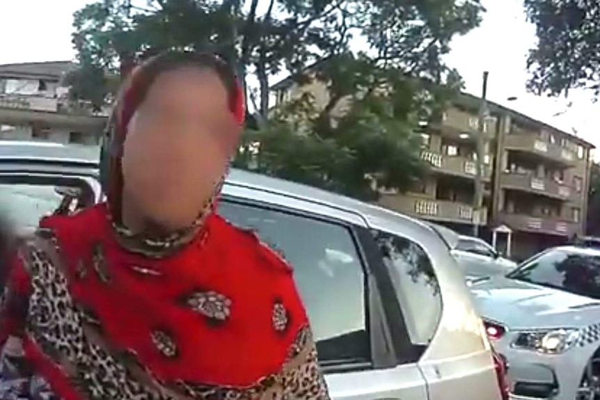 A woman wearing a headscarf stands in front of her car which has been pulled over by a police car.