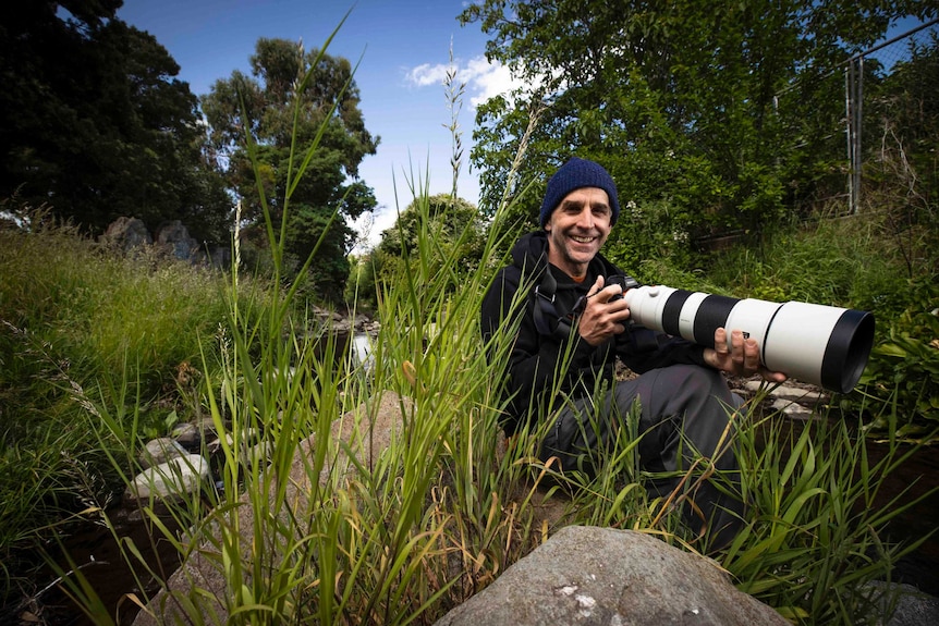 A man with a camera and a large lens sits on a river bank.