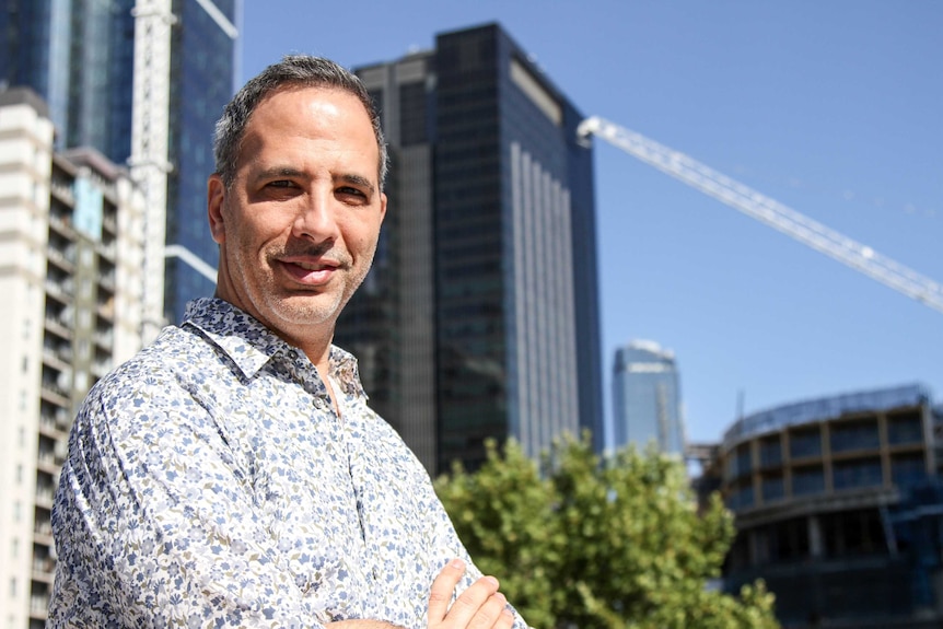A man with greying hair and a floral shirt stands outside in front of a city skyline.
