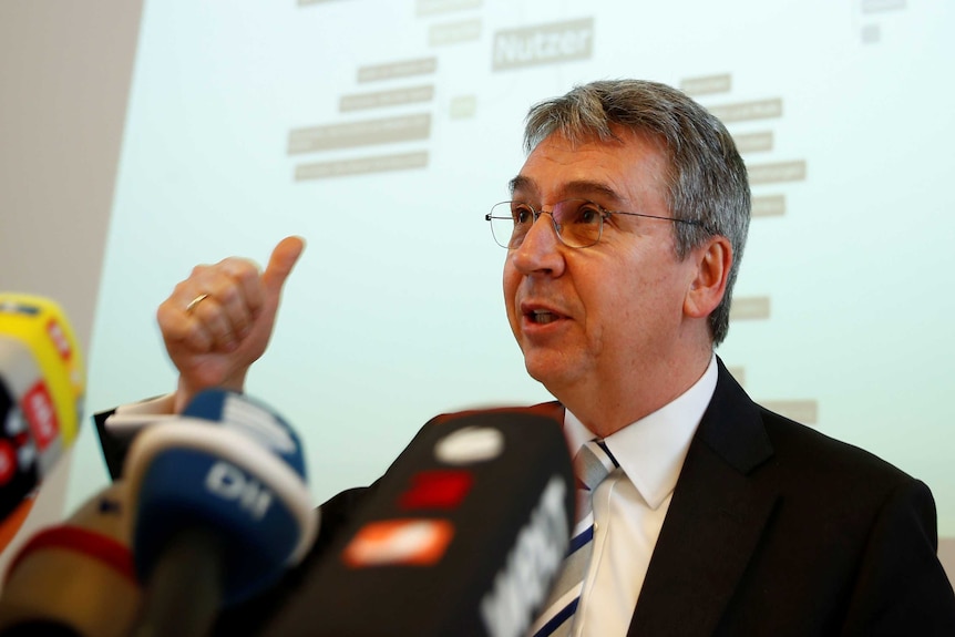 A caucasian man sitting down behind microphones points his right thumb back at a projector screen. He has wire-rimmed glasses.