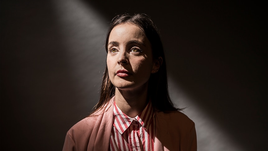 The performer stares out of frame, wearing a red striped blouse and a pink jacket. She stands against a shadowy wall