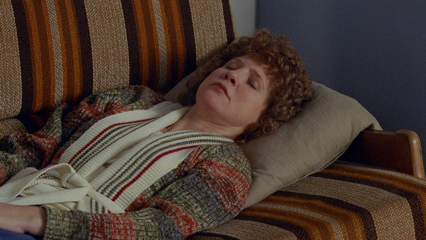 Woman with ginger perm and wearing 1970s-style cardigan lying with eyes closed on retro brown-striped couch.