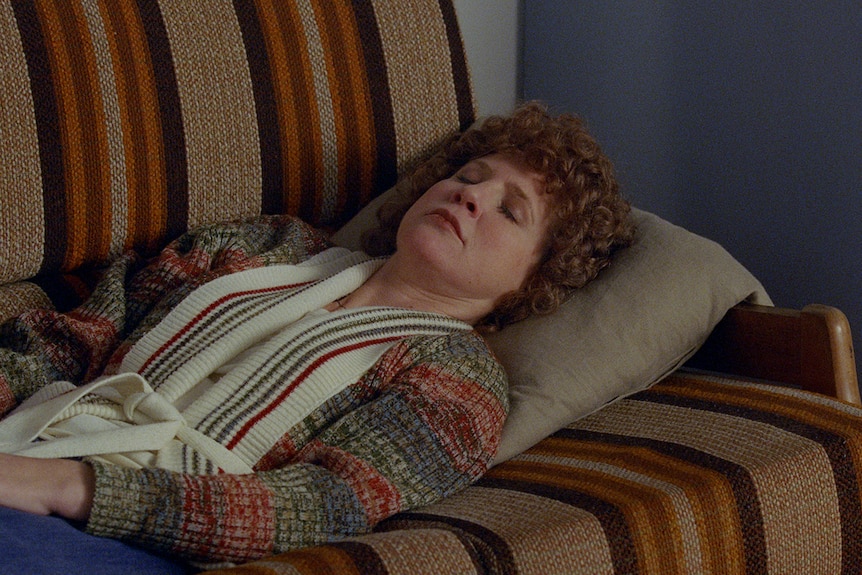 Woman with ginger perm and wearing 1970s-style cardigan lying with eyes closed on retro brown-striped couch.