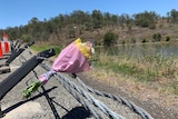 A bouquet of flowers with pink wrapping sits on wire fence next to a dam in a wooded country area.