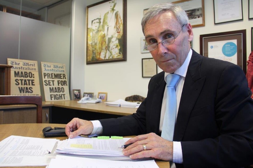 An older, silver-haired man in a dark suit sits at a desk in an office.