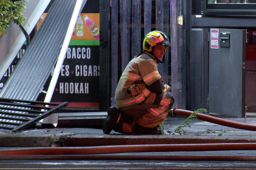 A firefighter wearing a reflective jacket and helmet kneels down beside a rose in front of a broken door of a shop.