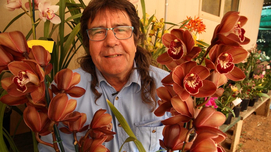 Stephen Lynch crouches and smiles between a tall, red orchid plant.