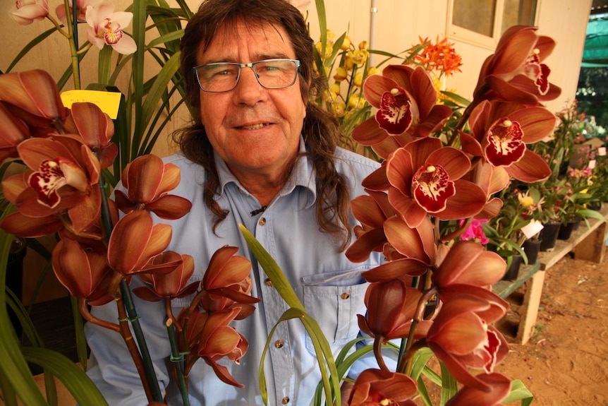 Stephen Lynch crouches and smiles between a tall, red and white orchid plant.